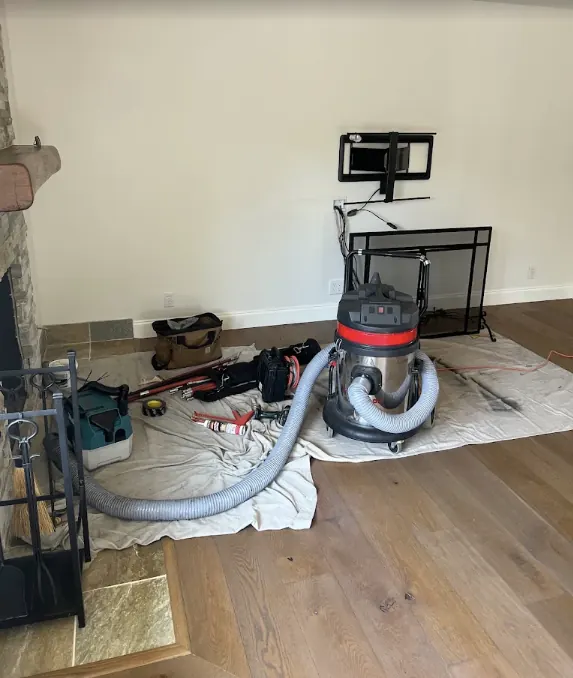 Chimney sweep tools and a vacuum laid out on drop cloths over a hardwood floor beside a stone fireplace