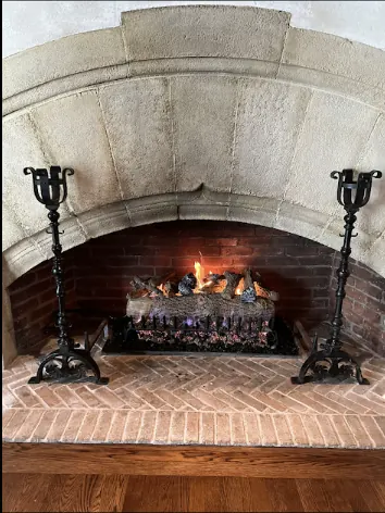 A gas log set burning in a carved stone arch fireplace with iron andirons