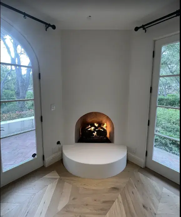 Gas logs burning in a small arched plaster fireplace with a rounded hearth, set between an arched glass door and French doors