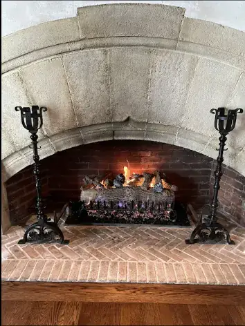 A gas log set burning in a carved stone arch fireplace with iron andirons
