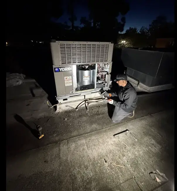 A technician kneeling at an opened York rooftop package unit at night, working by headlamp with the panel off