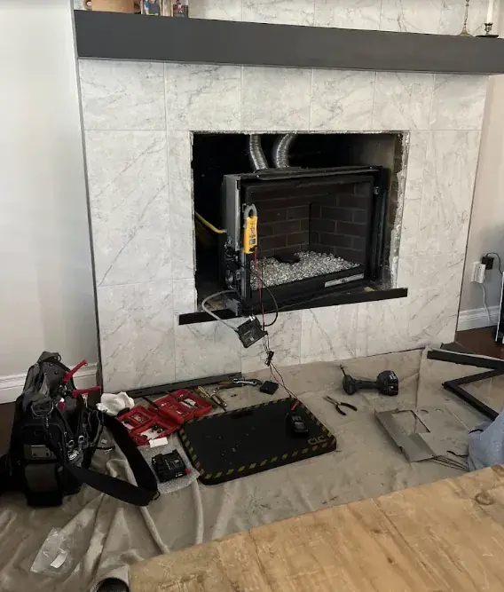 A gas fireplace insert being wired inside a marble tiled surround, with a multimeter clamped to the unit and tools laid out on a drop cloth