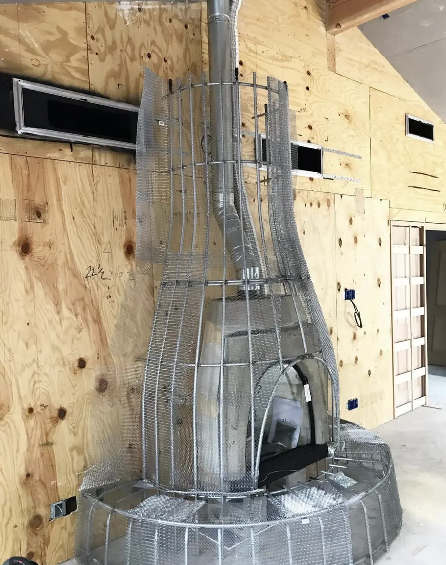 A kiva fireplace under construction: firebox and vent pipe wrapped in a hand shaped steel lath frame against plywood walls