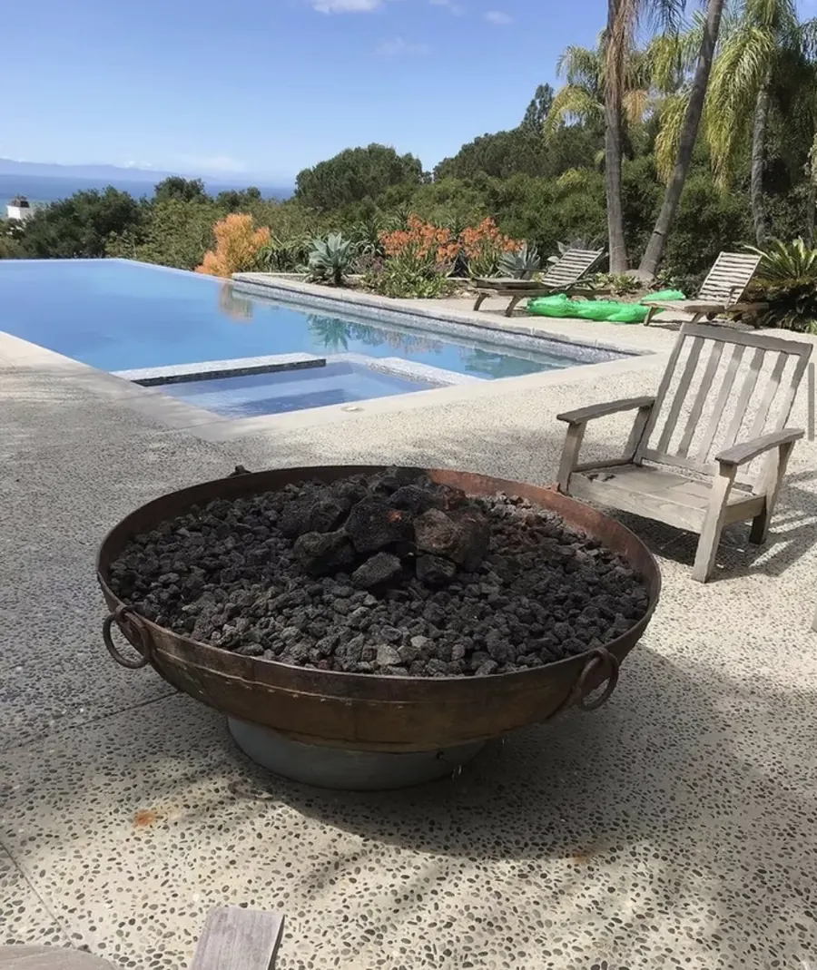 A steel fire bowl filled with lava rock beside a pool with the ocean beyond